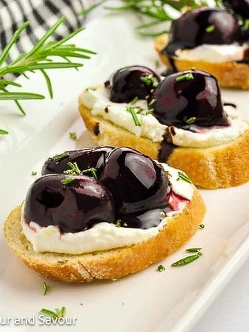 Three roasted cherry crostini with goat cheese on a white serving platter.