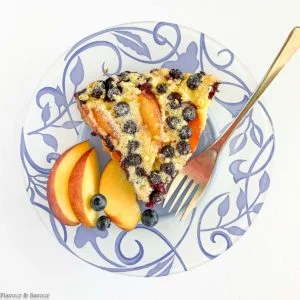 A slice of Gluten-Free Blueberry Peach Clafoutis on a blue patterned plate with fresh peach slices